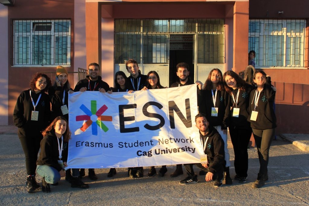 Yarının Kampı Ekibi ve Çağ Üniversitesi’nden gelen gönüllü öğrenciler okulun önünde ESN Cag University bayrağını tutuyor. Fotoğrafta 12 proje katılımcısı var. Ortalarında açtıkları bayrakla kameraya gülümsüyorlar.