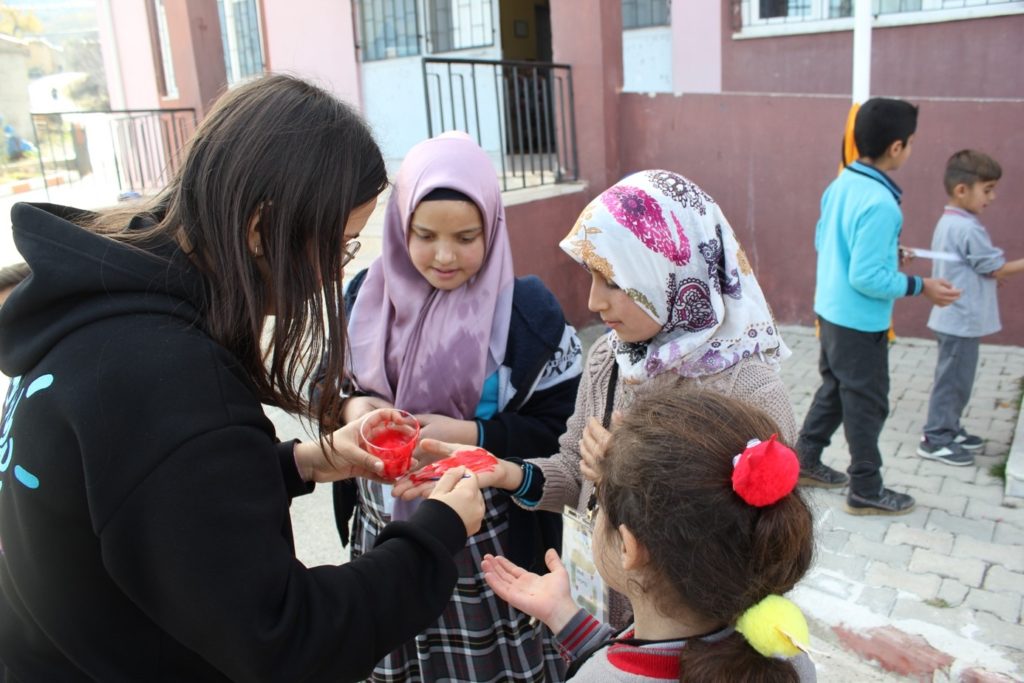 Fotoğrafta Yarının Kapmı Prorjesi gönüllüsü 2 kız öğrencinin ellerini Yarına İz Bırak! Sloganlı bayrakta el izi yapabilmeleri için kırmızı boyayla boyuyor. Okulun bahçesindeler ve bir kız öğrenci onları izliyor.