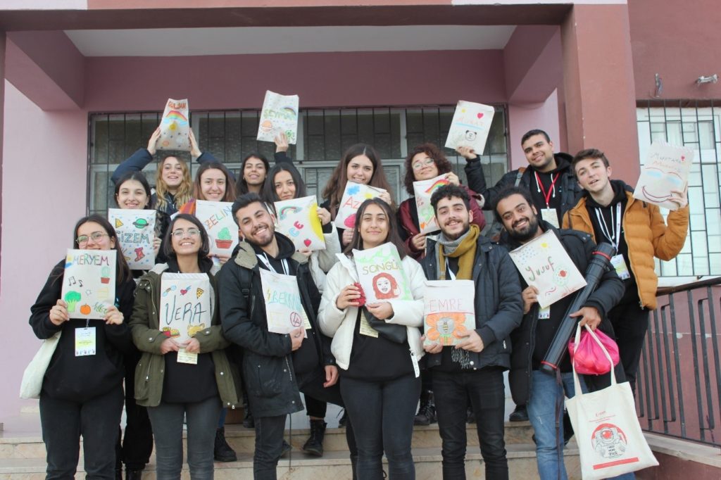 Fotoğrafta Yarının Kampı gönüllüleri gerçekleştirmiş oldukları zarf boyama çalışmalarıyla birlikte okulun önünde poz vermişlerdir. Zarfların üzerine kendi isimlerini içeren renkli bir yazı çalışmasının yanı sıra çiçeklere, ağaçlara, bulutlara ve hayvanlara kendi yapmış oldukları çizimlerle yer vermişler.