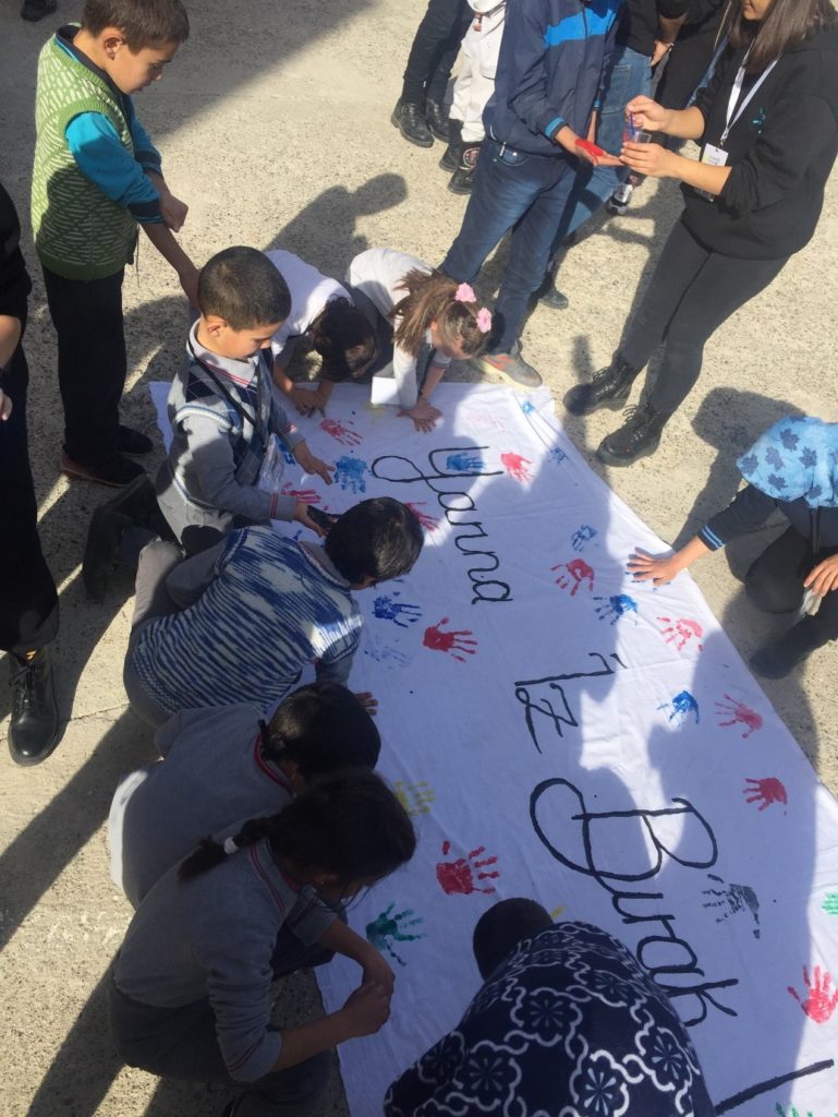 Fotoğrafta okulun bahçesinde Yerde duran ‘Yarına İz Bırak!’ sloganlı pankarta öğrencilerin el izi boyamalarını sarı, mavi, kırmızı, siyah, yeşil renkler ile gerçekleştirdiği gözlemlenmektedir. Yarının kampı ekibinden bir kişi de bir erkek öğrencinin elini kırmızıya boyuyor.